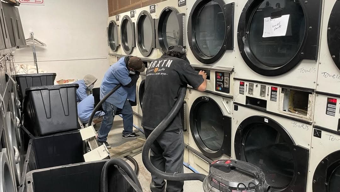 Workers cleaning commercial dryers in a laundromat with vacuum equipment.